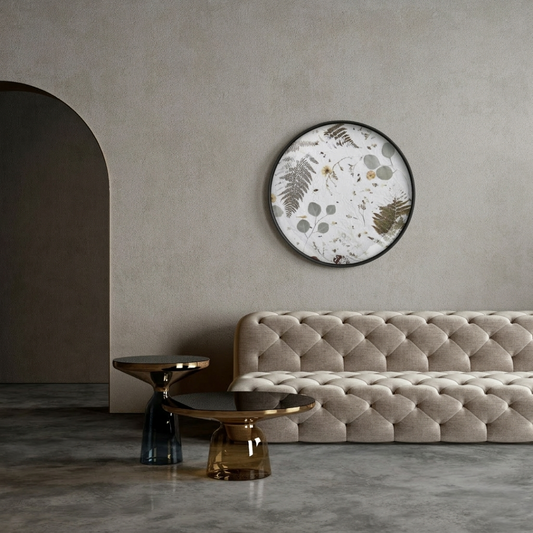 The Nudiflorum R55/06 botanical print hangs above a beige quilted velour sofa in a modern living room with walls painted in a warm shade of grey; the floor is concrete and there are two contemporary glass coffee tables