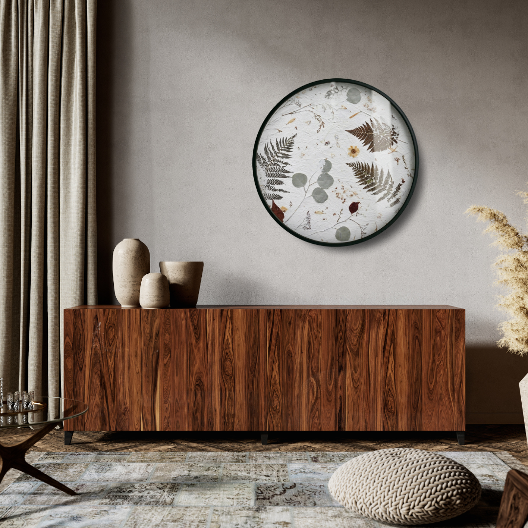 A round wall decoration with a floral motif, the Nudiflorum R55/09, hangs on a raw concrete wall above a modern wooden chest of drawers, next to a curtain in a neutral colour. Decorative ceramic vases sit on the chest of drawers.