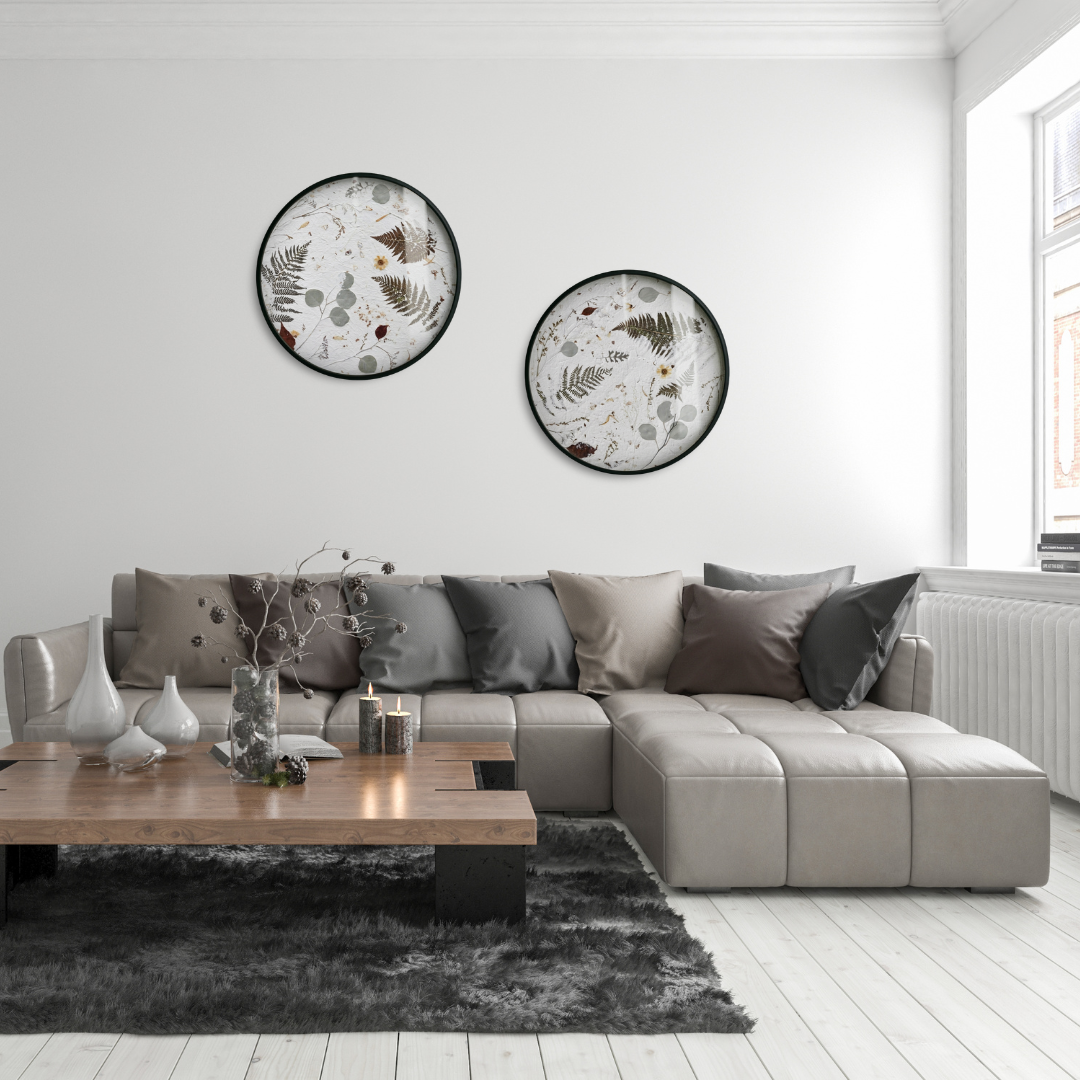 A Nudiflorum R55/09 botanical object (set of 2) in a modern, bright living room, hung above a grey leather sofa. White wooden flooring, a graphite-coloured rug, and on it a wooden coffee table with vases and candles. Lots of cushions on the sofa