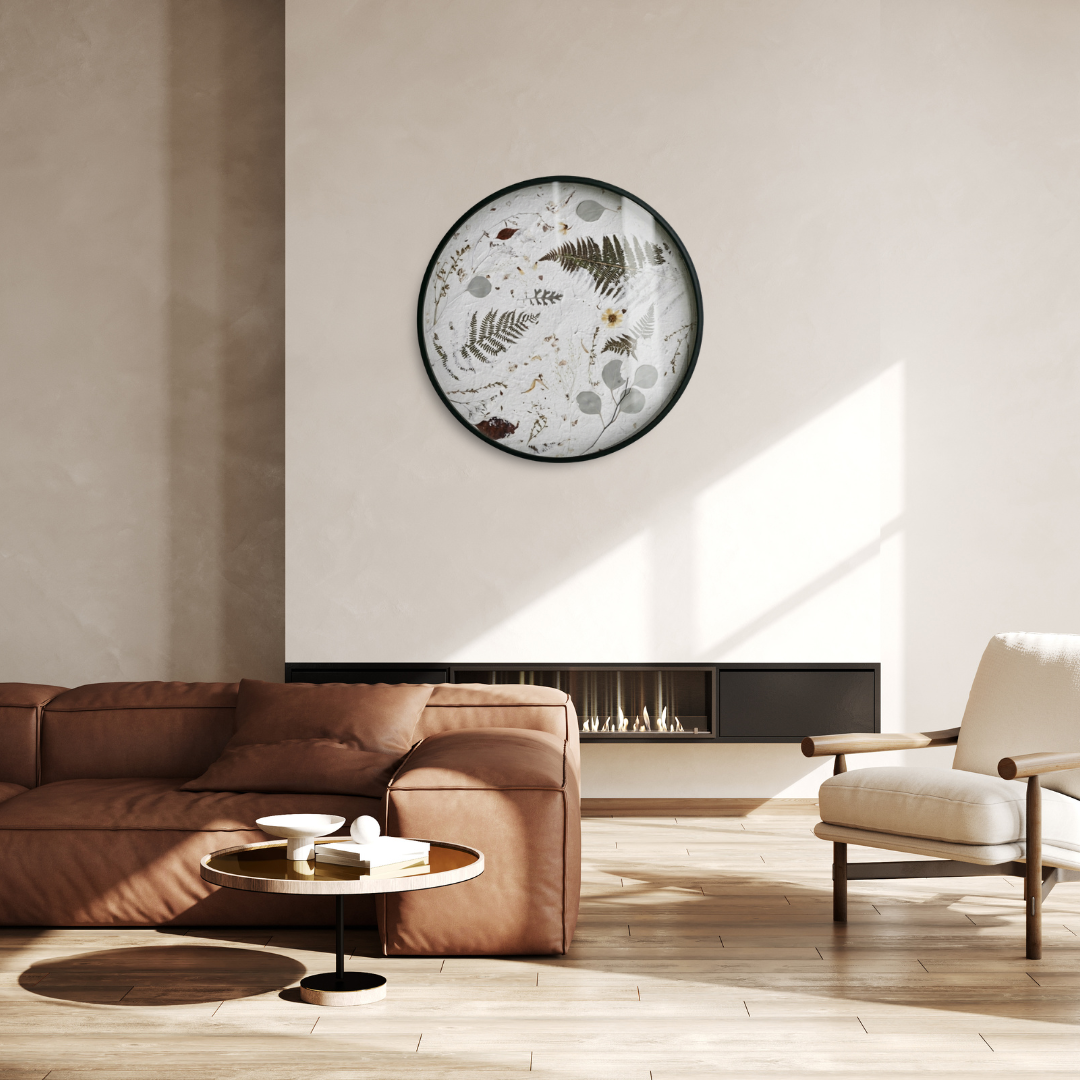 A round wall botanical object , the Nudiflorum R55/10, hangs on the wall above the fireplace in a minimalist living room featuring a caramel-coloured leather sofa and a wooden armchair upholstered in beige. Plenty of natural light